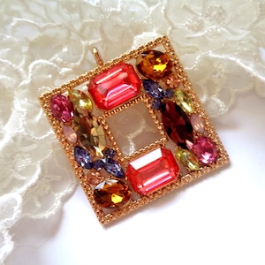 Könnte beinhalten: Ein quadratischer Anhänger mit goldfarbenem Rahmen, gefüllt mit verschiedenen bunten Edelsteinen. Die Edelsteine umfassen rote, orange, gelbe, violette und rosa Farbtöne. Der Anhänger liegt auf einem weißen Spitzengrund.