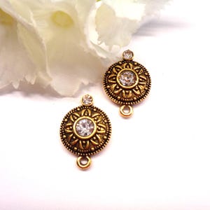 Peut inclure: Deux composants de bijoux de couleur or antique. Chaque composant présente un cristal clair central entouré d'un motif floral détaillé. Un cristal plus petit est placé au-dessus du motif principal, et une boucle est en bas pour la fixation.