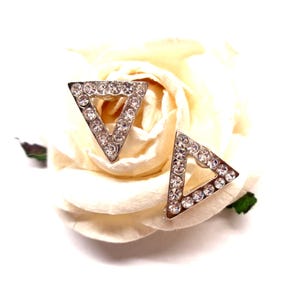 Peut inclure: Une paire de boucles d'oreilles triangulaires argentées, chacune bordée de petits cristaux transparents. Les boucles d'oreilles sont posées sur une rose blanche crémeuse, créant une présentation délicate et élégante.