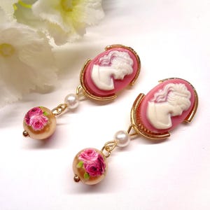 Peut inclure: Une paire de boucles d'oreilles pendantes avec un camée ovale rose et une silhouette de profil blanche dans un cadre doré. Une petite perle blanche relie le camée à une perle beige ronde ornée d'un motif floral de roses roses.