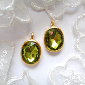 Peut inclure: Deux pierres précieuses ovales, facettées, vert olive, serties dans des montures en métal doré. Chaque gemme a une petite boucle en haut. Le fond est blanc avec de la dentelle délicate.