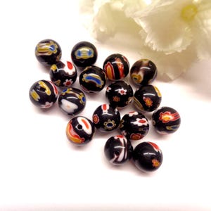 Peut inclure: Une collection de perles rondes en verre noir ornées de motifs millefiori complexes aux couleurs vives, notamment rouge, jaune, bleu et blanc. Chaque perle présente une surface lisse et polie avec un trou central, révélant des motifs floraux et abstraits uniques.