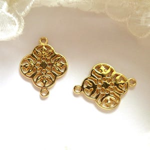 Peut inclure: Deux connecteurs en filigrane dorés avec un motif floral à quatre pétales. Chaque connecteur comporte trois petites boucles pour la fixation. Les connecteurs sont sur une surface blanche avec une bordure en dentelle.