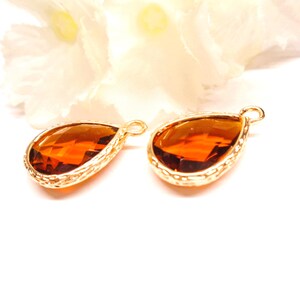 Peut inclure: Deux pendentifs en forme de goutte d'eau, de couleur ambre, avec une bordure en filigrane dorée. Les pierres à facettes sont posées sur un fond blanc, avec une fleur blanche floue dans le coin supérieur droit. Chaque pendentif a une petite boucle en haut.
