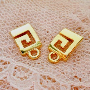 Könnte beinhalten: Zwei goldfarbene, quadratische Schmuckbeschläge mit einem griechischen Schlüsseldesign. Jeder Beschlag hat unten eine kleine Schlaufe zur Befestigung an anderen Komponenten. Die Beschläge werden auf einer rosa Spitzenoberfläche präsentiert.