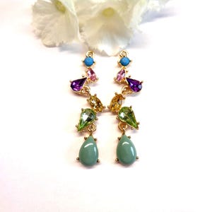 Peut inclure: Une paire de boucles d'oreilles dorées avec des pierres précieuses colorées. Les boucles d'oreilles présentent des pierres turquoise, roses, violettes, jaunes et vertes de différentes formes, dont des gouttes. Les boucles d'oreilles sont posées sur un fond blanc avec une fleur blanche.
