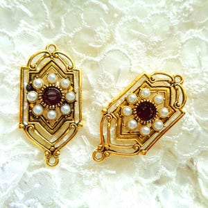 Puede incluir: Dos colgantes decorativos dorados. Cada uno presenta una piedra central redonda, de color rojo oscuro, rodeada por un anillo de pequeñas perlas blancas. Los colgantes tienen un diseño calado con bordes decorativos y un lazo en la parte superior.