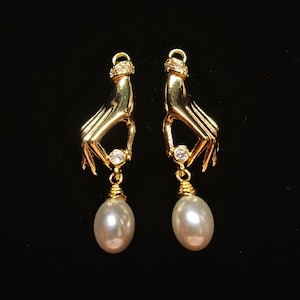 Peut inclure: Une paire de boucles d'oreilles dorées représentant une main tenant une petite pierre transparente entre les doigts, avec une perle blanche ovale suspendue. Chaque boucle possède une pierre transparente au poignet et une attache en haut.