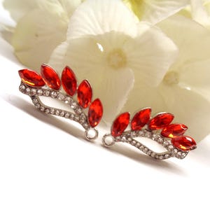Puede incluir: Un par de pendientes trepadores de metal plateado con un diseño curvo, adornados con cristales rojos de talla marquesa y una hilera de pequeños diamantes de imitación redondos y transparentes. Cada pieza incluye una pequeña anilla circular.