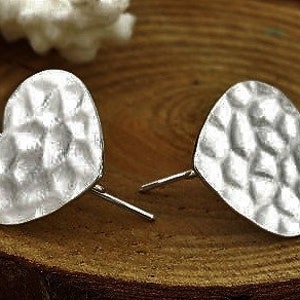 Peut inclure: Une paire de boucles d'oreilles argentées en forme de cœur avec une texture martelée. Les boucles d'oreilles sont présentées sur une surface en bois, mettant en valeur leur design. La forme de cœur ajoute une touche romantique.