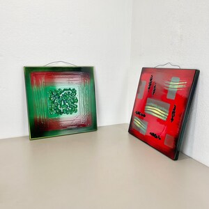 Könnte beinhalten: Zwei quadratische dekorative Wandkunstwerke. Eines ist grün und rot mit einem strukturierten, blütenartigen Design. Das andere ist rot mit grauen und gelben abstrakten Formen. Beide haben schwarze Rahmen und Aufhängedrähte.