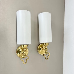 Puede incluir: Un par de apliques de pared de latón con pantallas de vidrio blanco esmerilado. Cada aplique tiene un detalle decorativo en forma de llave.