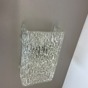 May include: A clear glass wall sconce with a textured, rippled surface. The sconce has a rectangular shape and is designed to be mounted on a wall.