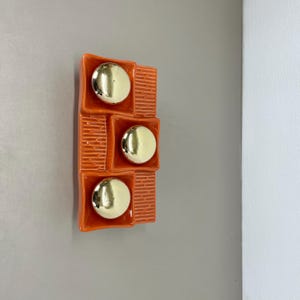 Puede incluir: Arte de pared de cerámica naranja con tres círculos dorados en un patrón de cuadrícula.