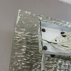 May include: Close-up of the back of a white ceiling light fixture with exposed wiring. The fixture is attached to a textured glass panel.