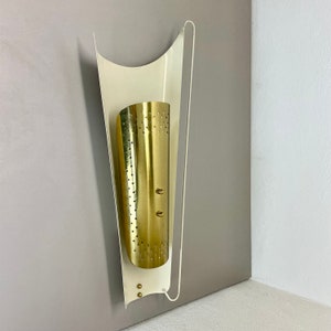 Könnte beinhalten: Eine weiße und goldene Wandleuchte mit einem perforierten Messing-Schirm. Die Leuchte hat ein modernes Design und ist perfekt, um jedem Raum einen Hauch von Stil zu verleihen.