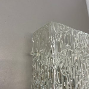 May include: A clear glass wall sconce with a textured, rippled surface. The sconce is square shaped and designed to be mounted on a wall.