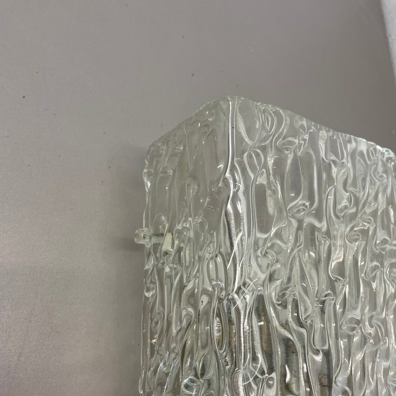 May include: A clear glass wall sconce with a textured, rippled surface. The sconce is square shaped and designed to be mounted on a wall.