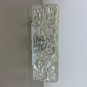 May include: A clear glass wall sconce with a textured surface. The sconce is mounted on a white wall.