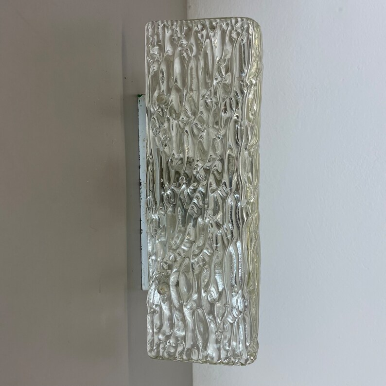 May include: A clear glass wall sconce with a textured surface. The sconce is mounted on a white wall.