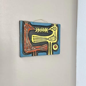 Könnte beinhalten: Ein quadratisches Keramik-Wandbild mit blauem Rand und schwarzem Innenraum. Das Kunstwerk zeigt zwei stilisierte Vögel in Rot und Gelb, mit einem fischartigen Element dazwischen. Das Stück wird an einer Schnur aufgehängt.