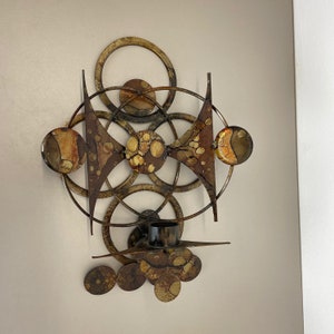Könnte beinhalten: Ein Wand-Kerzenhalter mit geometrischem Design. Der Halter ist aus Metall und hat eine kreisförmige Form mit mehreren sich überlappenden Kreisen. Die Kreise sind mit einem braunen und goldenen Farbmuster verziert. Der Halter hat einen einzelnen Kerzenhalter in der Mitte.