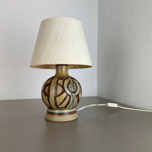 Könnte beinhalten: Eine Keramik-Tischlampe mit einem weißen Leinen-Lampenschirm. Die Lampenbasis hat ein geometrisches Design mit braunen, gelben und blauen Akzenten.
