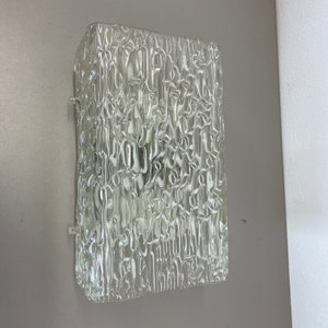 May include: A clear glass wall sconce with a textured surface. The sconce is square shaped and is mounted on a wall.
