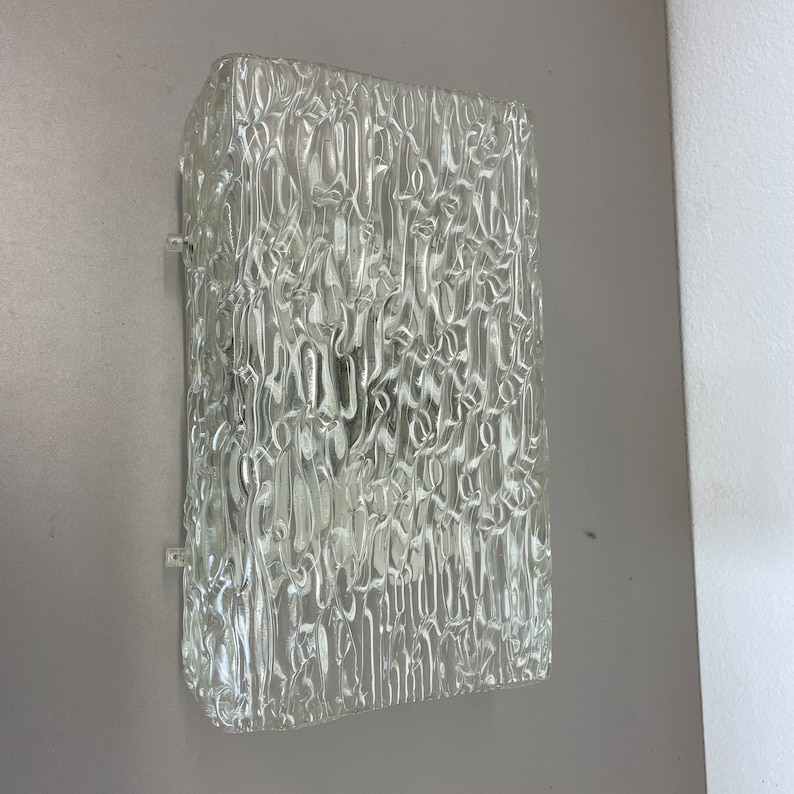 May include: A clear glass wall sconce with a textured surface. The sconce is square shaped and is mounted on a wall.