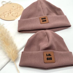 Könnte beinhalten: Zwei rosafarbene gerippte Strickmützen mit braunen Lederpatches. Die größere Mütze hat einen Patch mit der Aufschrift "BIG SIS" und die kleinere Mütze hat einen Patch mit der Aufschrift "LIL SIS".