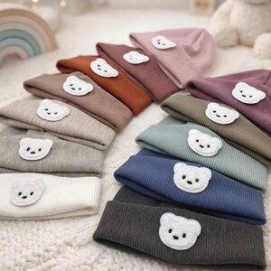 Könnte beinhalten: Eine Auswahl an gerippten Strickmützen in verschiedenen Farben, darunter Rosa, Braun und Blau. Jede Mütze hat eine weiße Bärenapplikation. Die Mützen sind auf einer weichen, weißen Oberfläche angeordnet.