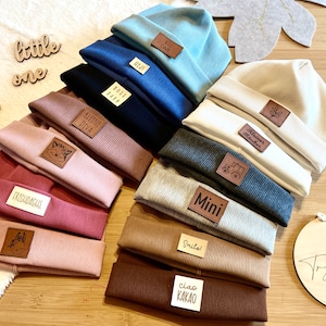 Könnte beinhalten: Eine Sammlung von Rippstrick-Beanies in verschiedenen Farben, darunter Blau, Rosa und Braun. Jede Beanie hat einen kleinen Lederaufnäher mit Text oder Grafik. Die Beanies sind auf einer Holzoberfläche angeordnet, mit einem Holzschild "Try Pinky".