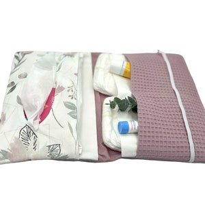 K&ouml;nnte beinhalten: Eine rosa-wei&szlig;e Wickeltasche mit Blumenmuster. Die Tasche ist ge&ouml;ffnet und enth&auml;lt Windeln, Feuchtt&uuml;cher, eine kleine Flasche Lotion und eine kleine Tube Salbe.