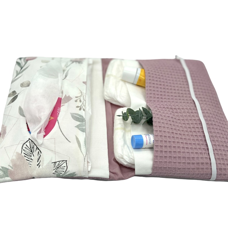 K&ouml;nnte beinhalten: Eine rosa-wei&szlig;e Wickeltasche mit Blumenmuster. Die Tasche ist ge&ouml;ffnet und enth&auml;lt Windeln, Feuchtt&uuml;cher, eine kleine Flasche Lotion und eine kleine Tube Salbe.