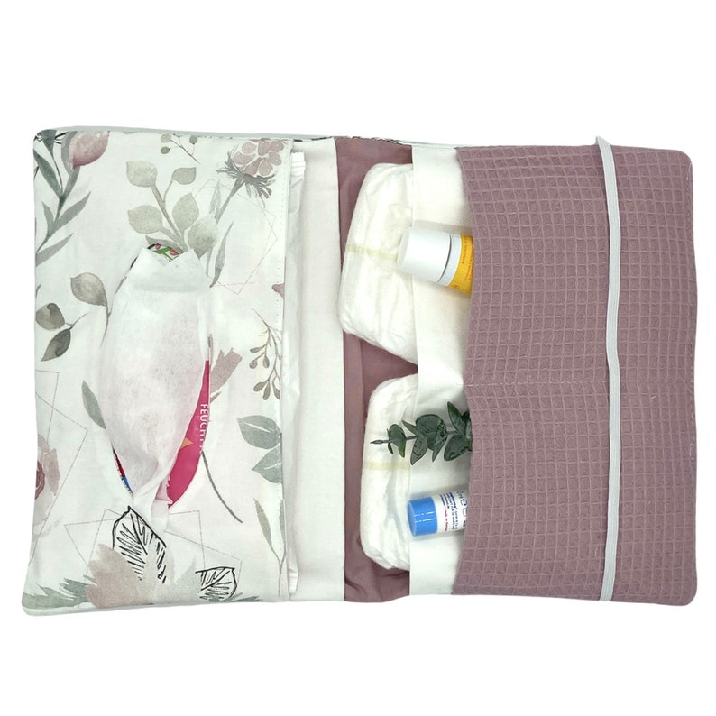 K&ouml;nnte beinhalten: Eine Wickeltasche mit floralem Druck in Rosa und Wei&szlig; mit Rei&szlig;verschluss. Die Tasche ist ge&ouml;ffnet und enth&auml;lt Windeln, Feuchtt&uuml;cher und Babylotion.