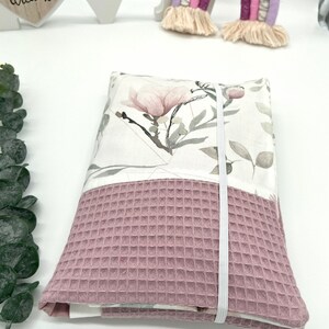 K&ouml;nnte beinhalten: Eine rosa-wei&szlig;e Stofftasche mit Blumenmuster und einem wei&szlig;en Gummibandverschluss. Die Tasche ist gefaltet und liegt auf einer wei&szlig;en Oberfl&auml;che.