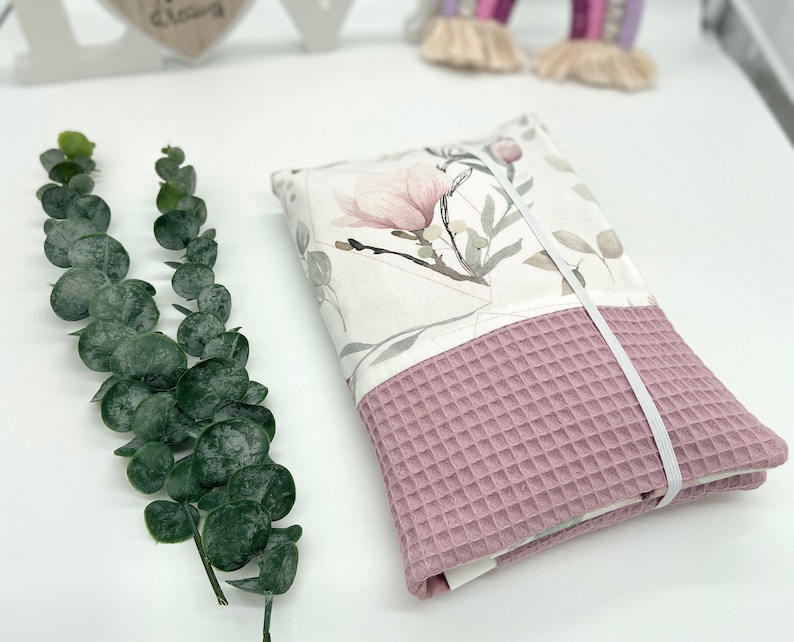 K&ouml;nnte beinhalten: Ein rosa-wei&szlig;es Stoff-Spucktuch f&uuml;r Babys mit Blumenmuster und einem wei&szlig;en Gummiband. Das Tuch ist gefaltet und mit dem Gummiband gebunden.