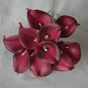 Könnte beinhalten: Ein Strauß burgunderfarbener Calla-Lilien mit weißen Zentren. Die Lilien sind in einer Gruppe angeordnet und haben eine samtige Textur.