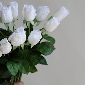 Op de afbeelding: Een boeket witte rozen met groene bladeren. De rozen zijn gerangschikt in een handgehouden boeket.