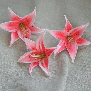 Puede incluir: Tres flores de lirio artificiales de color rosa con manchas de color rosa oscuro sobre un fondo de tela gris claro.