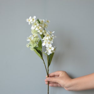 May include: A single stem of white artificial flowers with green leaves. The flowers are clustered together and have a delicate appearance.