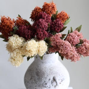 Può includere: Un vaso bianco strutturato pieno di fiori finti colorati. La composizione comprende fiori arancioni, bordeaux, crema e rosa con foglie verdi. Il vaso è posto su una superficie bianca su uno sfondo semplice.