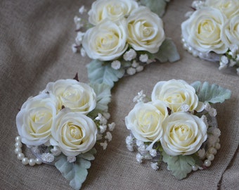 Puede incluir: Cinco boutonnières de rosas blancas con detalles de perlas y vegetación. Cada boutonnière tiene tres rosas blancas y pequeñas flores blancas.