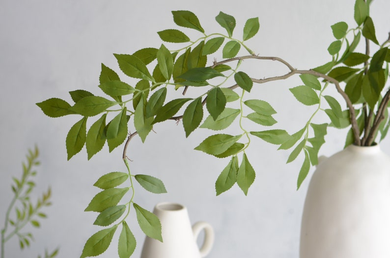 May include: A single branch of artificial greenery with green leaves in a white ceramic vase. The leaves are arranged in a cascading pattern, creating a natural and elegant look.