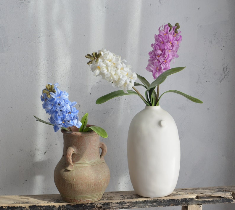 May include: A still life featuring three hyacinth flower arrangements. One arrangement has blue flowers in a rustic brown vase. Another has white flowers, and the third has purple flowers, both in a white, modern vase. The background is a neutral gray wall.