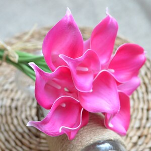 Puede incluir: Un ramo de vibrantes calas rosas con bordes blancos. Las flores est&aacute;n dispuestas en un jarr&oacute;n de vidrio transparente envuelto con hilo. El ramo descansa sobre una superficie de mimbre tejido, creando una exhibici&oacute;n natural y elegante.