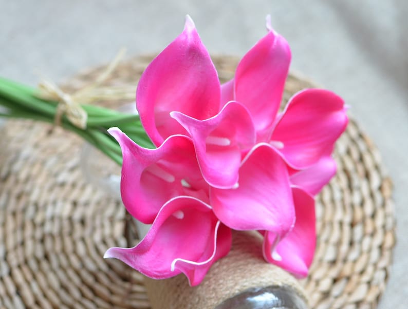 Puede incluir: Un ramo de vibrantes calas rosas con bordes blancos. Las flores est&aacute;n dispuestas en un jarr&oacute;n de vidrio transparente envuelto con hilo. El ramo descansa sobre una superficie de mimbre tejido, creando una exhibici&oacute;n natural y elegante.