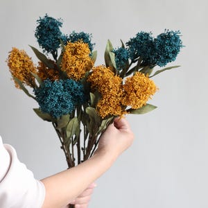 May include: A bouquet of artificial flowers featuring teal and golden yellow clusters. The faux flowers have green leaves and brown stems. The bouquet is held by a person wearing a white shirt against a light gray background.