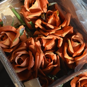 May include: A box of artificial orange roses. The roses are arranged in a bouquet and are in a clear plastic bag. The roses are in a cardboard box with a brown corrugated cardboard liner.