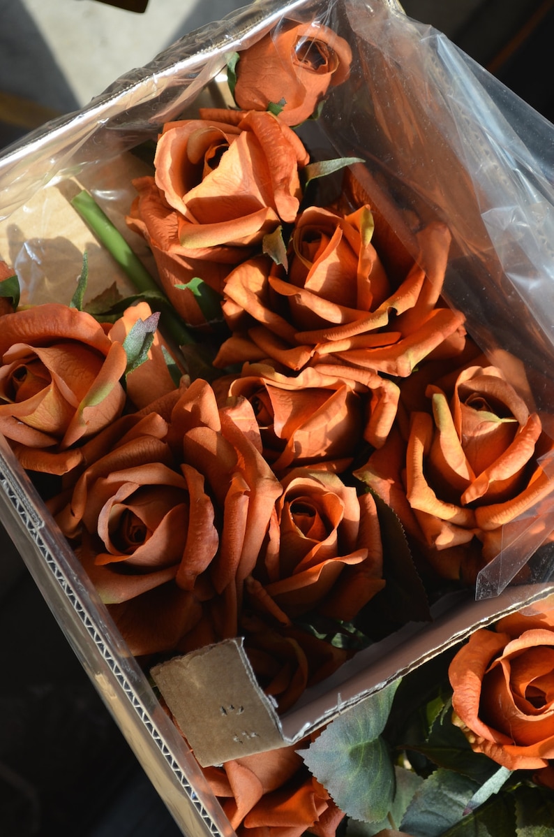 May include: A box of artificial orange roses. The roses are arranged in a bouquet and are in a clear plastic bag. The roses are in a cardboard box with a brown corrugated cardboard liner.